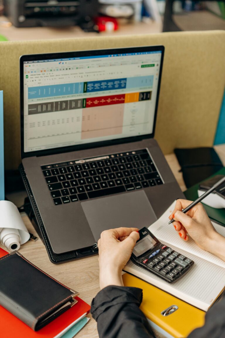 Hands writing notes beside a laptop displaying data in a bright office setup.